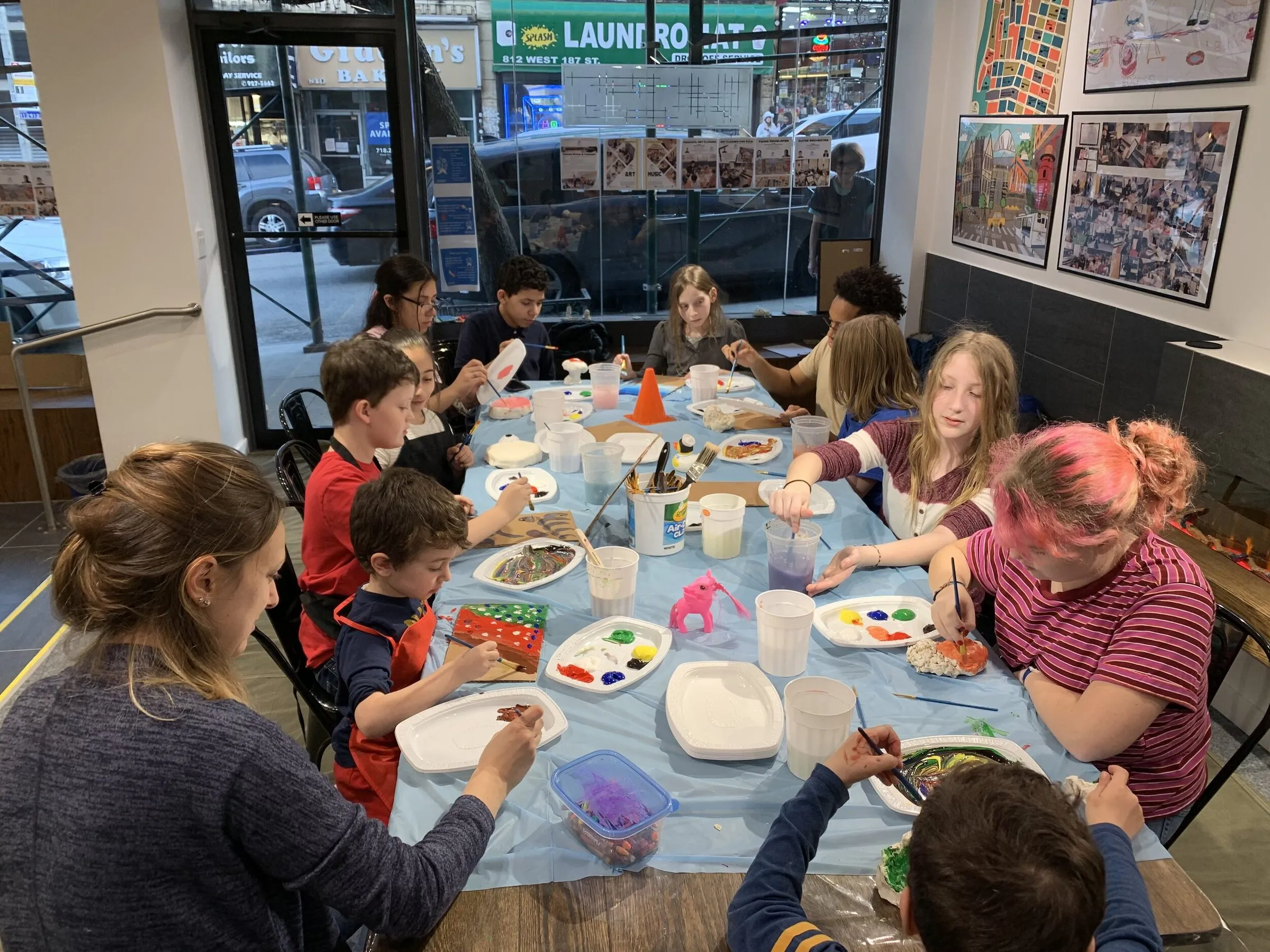 Children paint at a long table covered in paper, with framed student art on the wall and the street visible through the storefront windows.