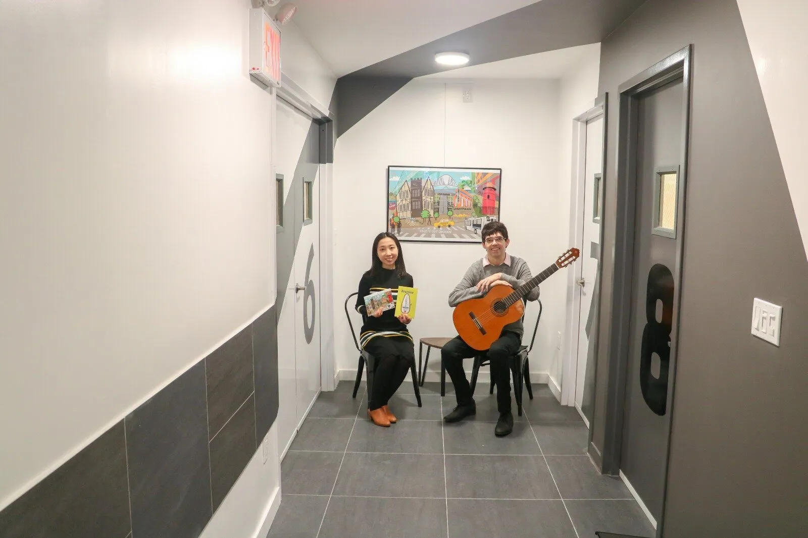 Two musicians, one holding a guitar, sit in a gray hallway lined with framed artwork beside numbered studio doors.