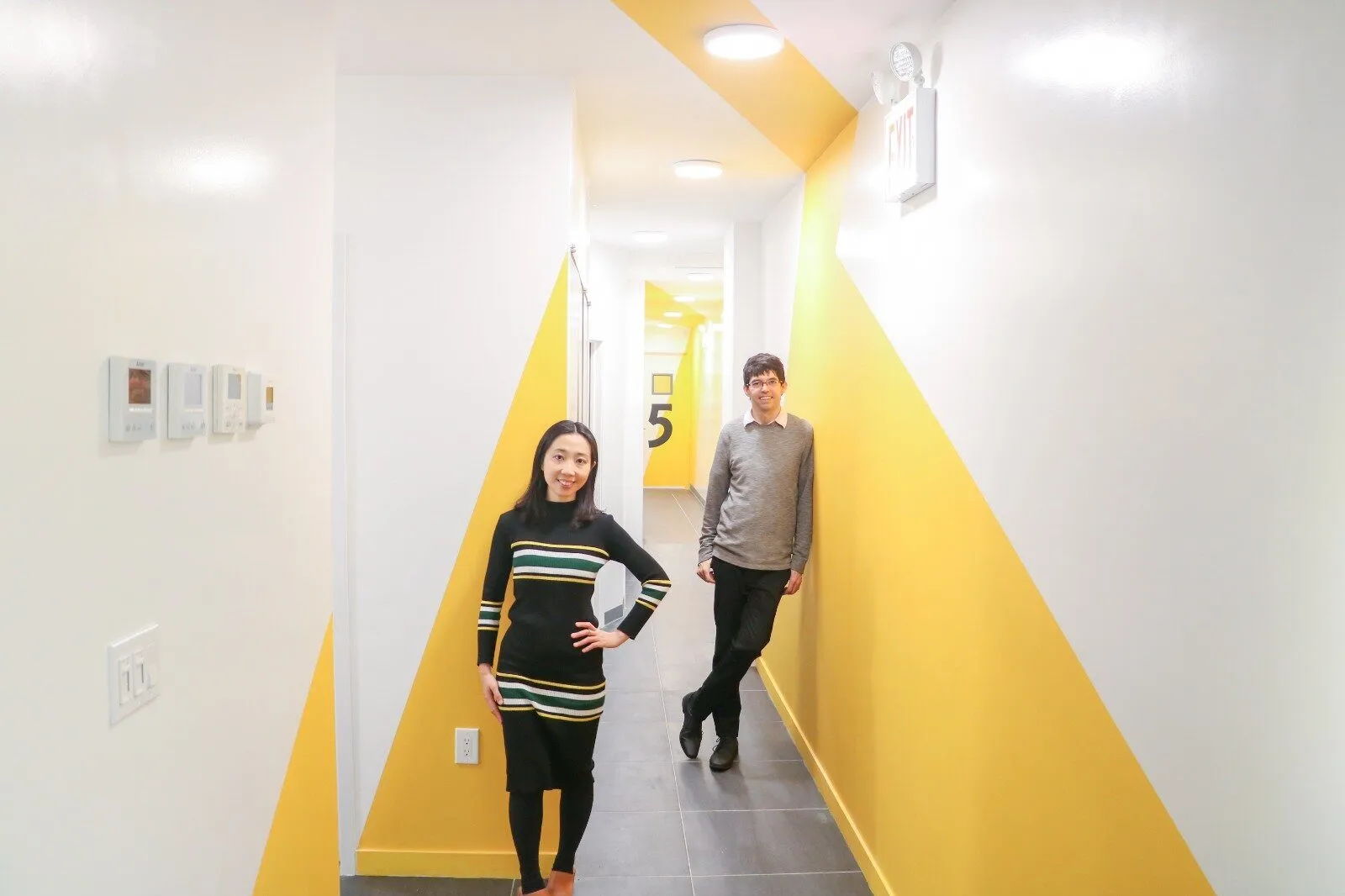A white corridor with a large yellow triangle painted across the floor-to-ceiling wall; two people stand along the hallway.