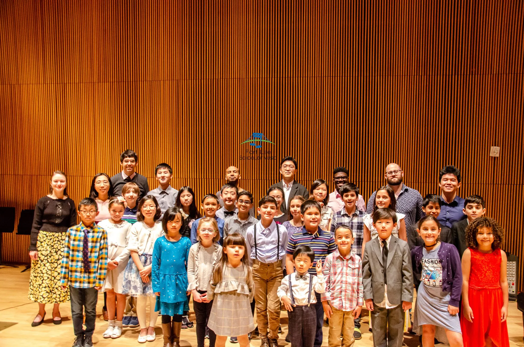Dozens of students in concert dress pose on a wood-paneled stage at the DiMenna Center for Classical Music.