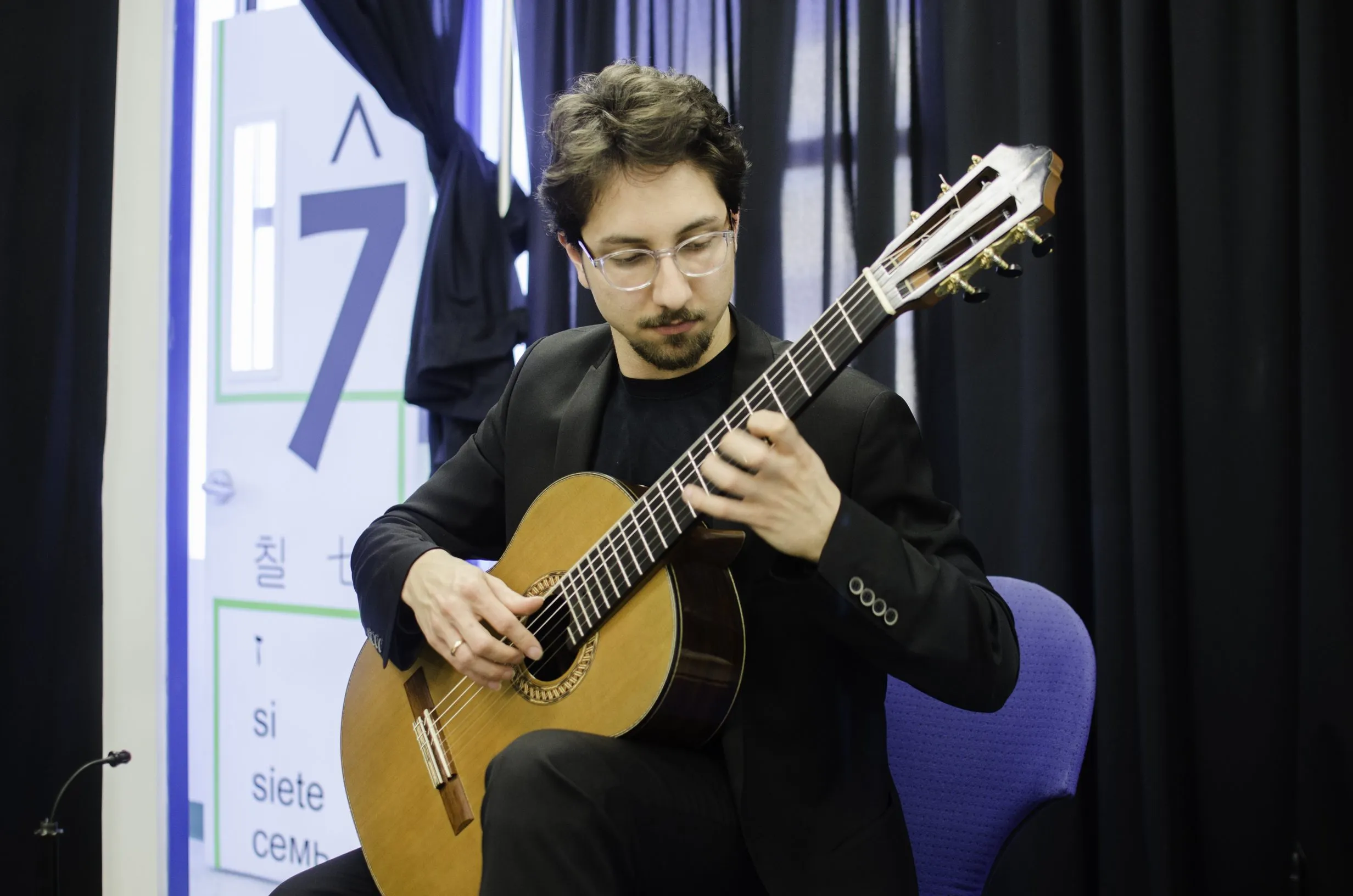 A classical guitarist performs in front of a studio door painted with the numeral 7, its solfège syllable 'si', and translations.