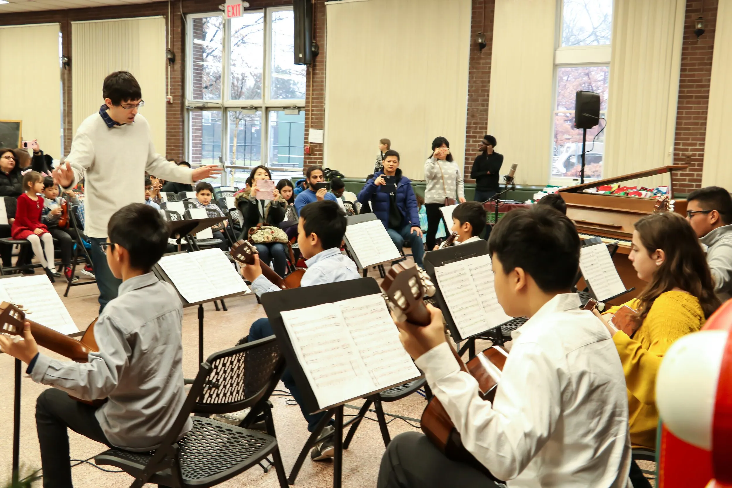 A teacher conducts a children's classical guitar orchestra seated at music stands in a sunlit hall.