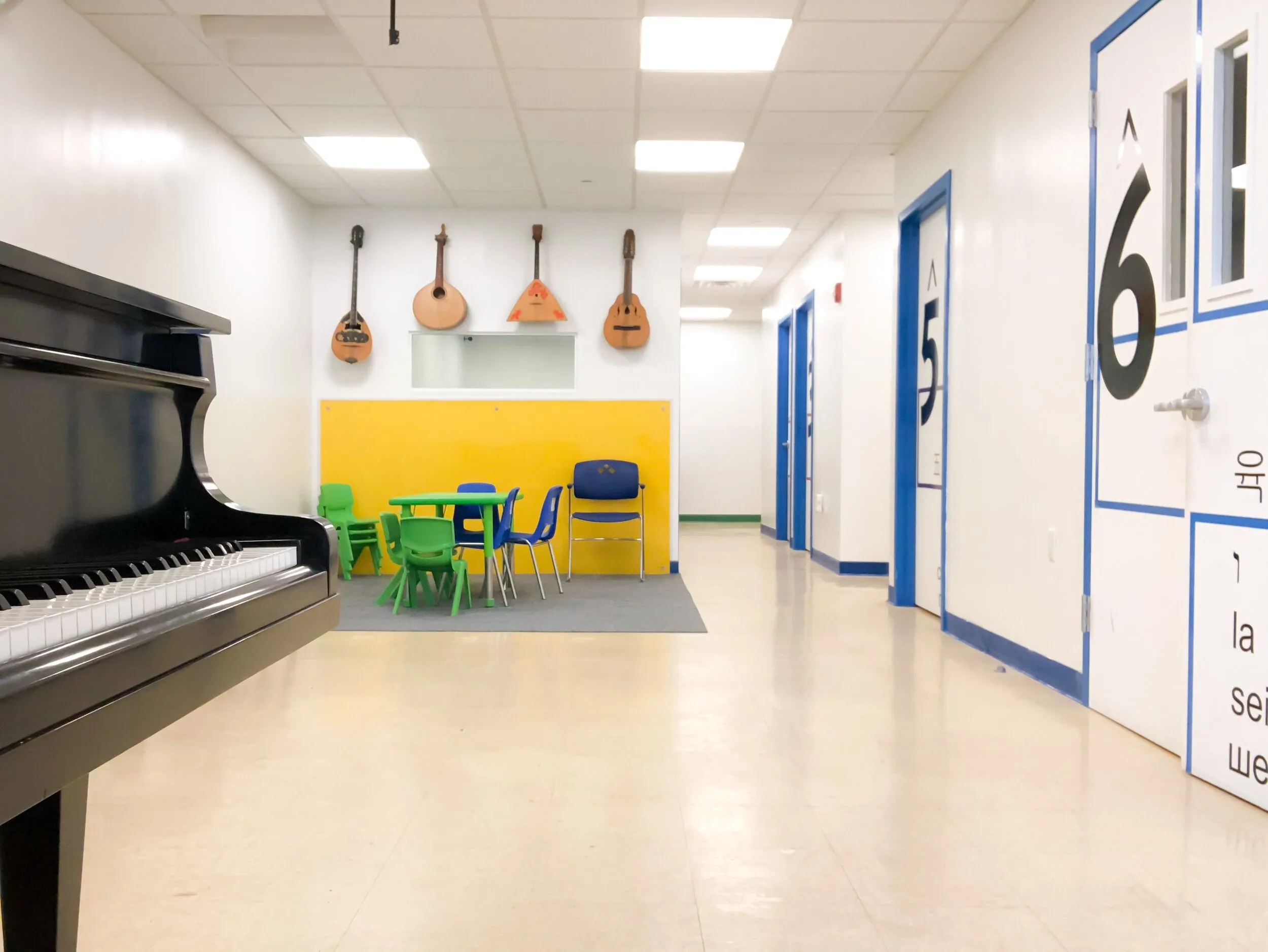 A bright hallway with a black grand piano, a yellow accent wall with children's tables, a wall of hanging folk instruments, and blue-framed doors marked with large numerals.