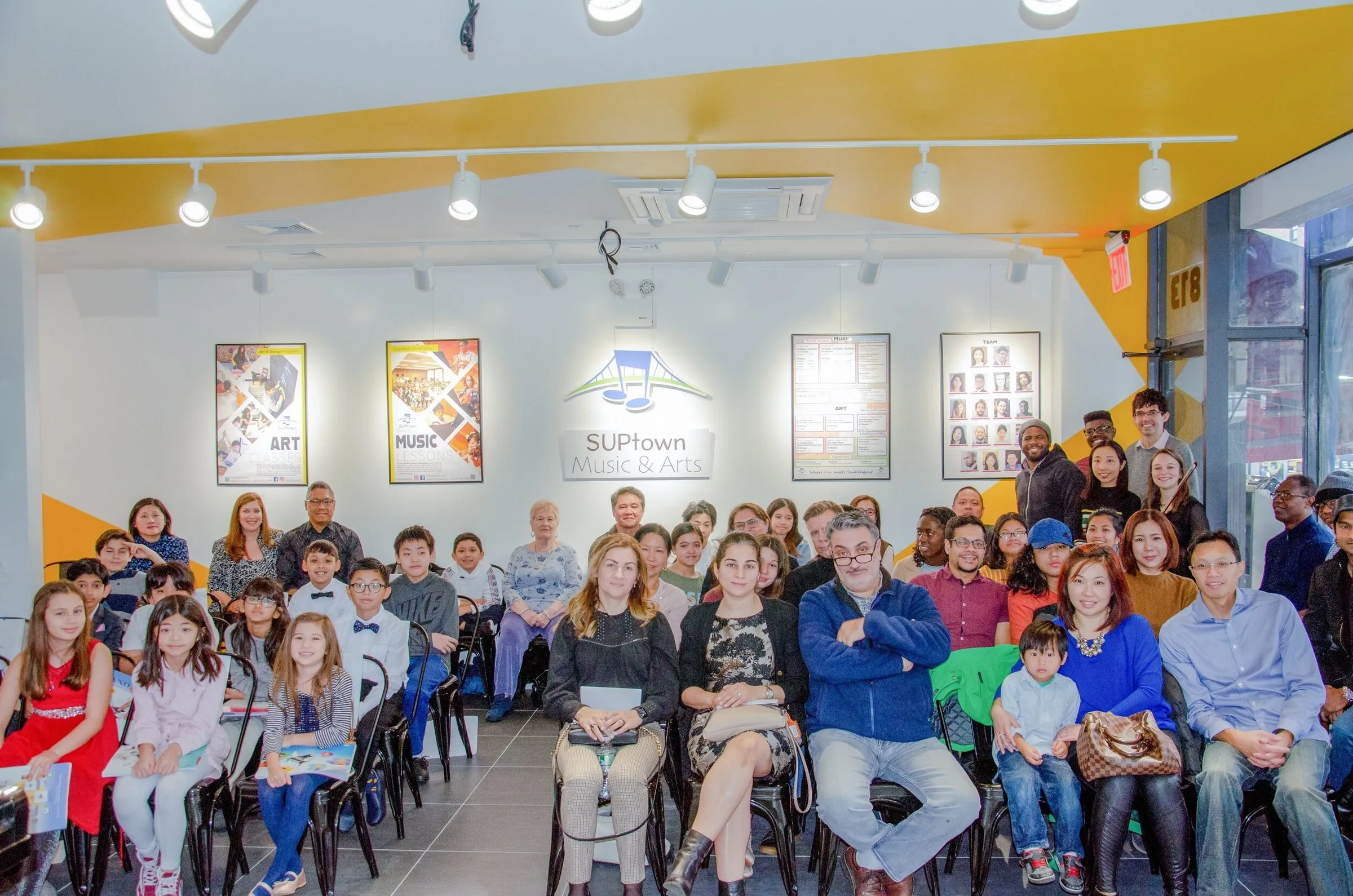 A full audience of families and students seated in rows in the SUPtown gallery, framed posters and the SUPtown logo on the wall behind them.