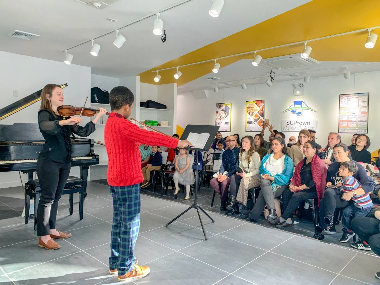 A violinist and a young student in a red sweater perform side by side in front of a full room, with the SUPtown logo on the wall behind the audience.