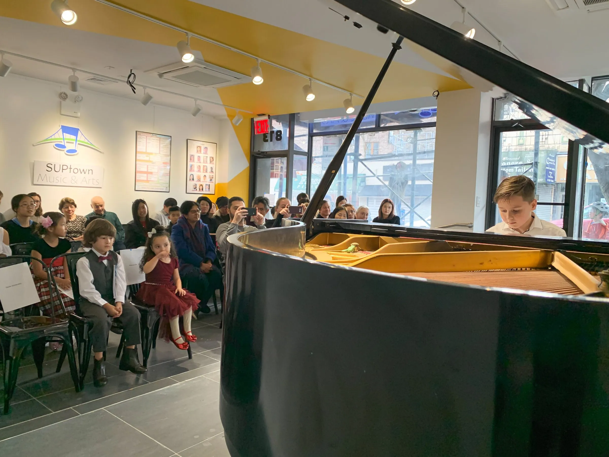 A grand piano in the foreground of the gallery room as an audience of families watches a student performance.