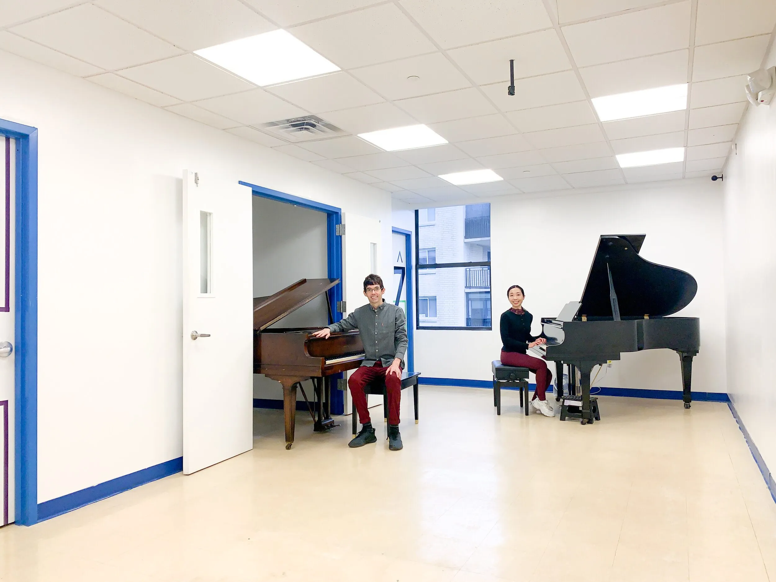 A large white studio with two grand pianos and a player seated at each, with blue trim outlining the doors and floor.