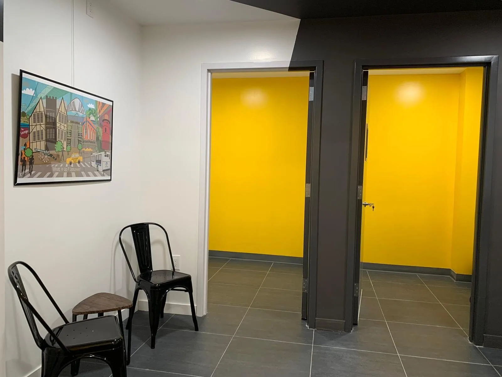 Two bright yellow practice-room doorways set into a charcoal wall, with framed artwork and black metal chairs.