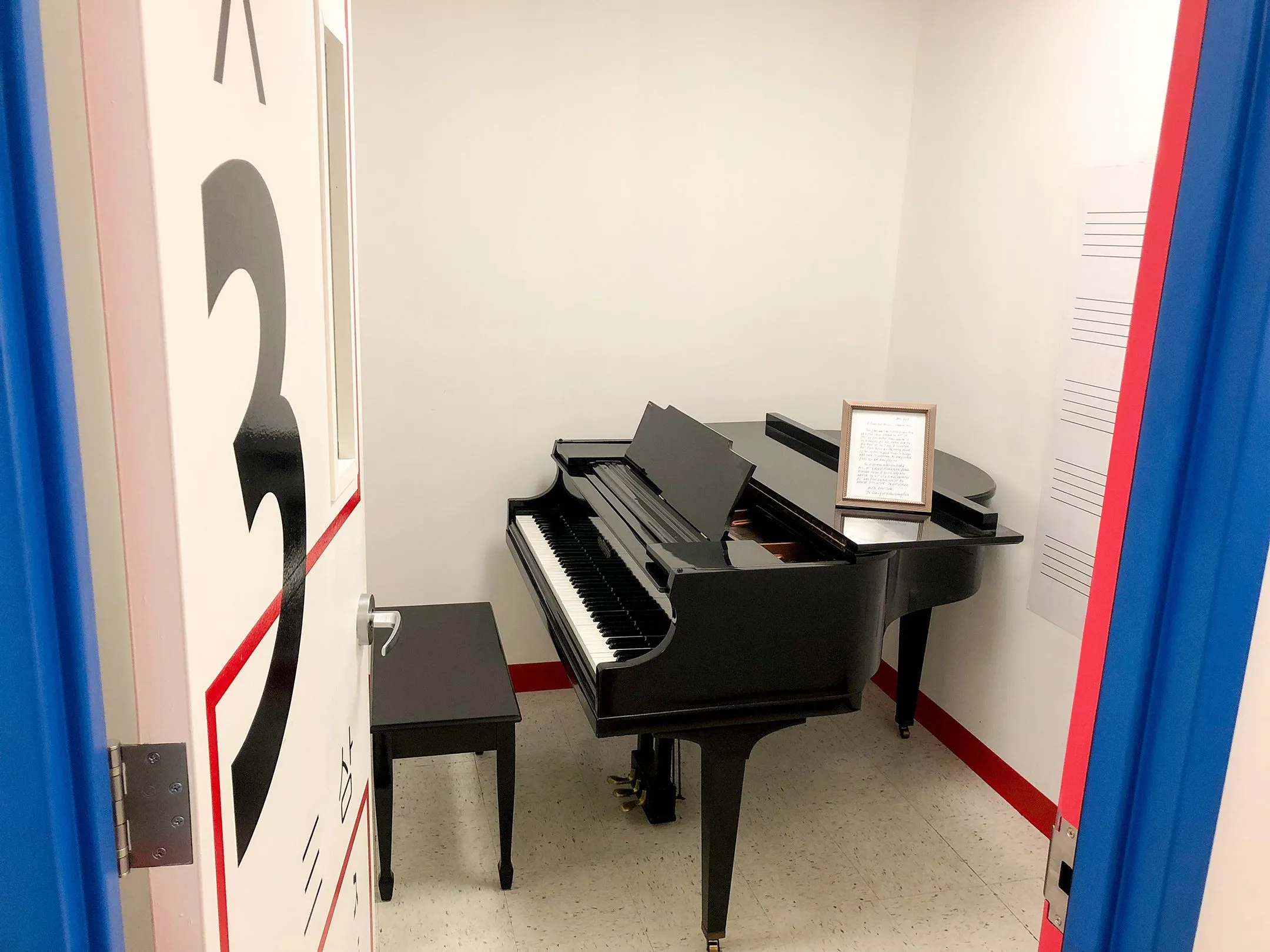 An open studio door painted with a huge numeral 3 reveals a small room with a black baby grand piano, framed by red and blue trim.