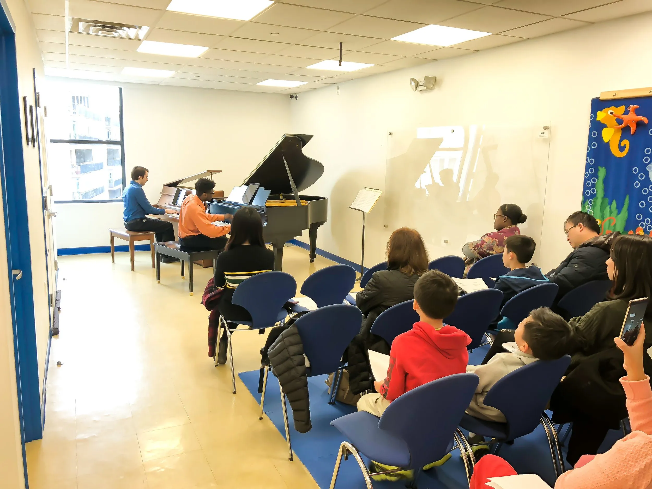 An audience in blue chairs watches a piano and violin performance in a studio with blue curtains and a grand piano.