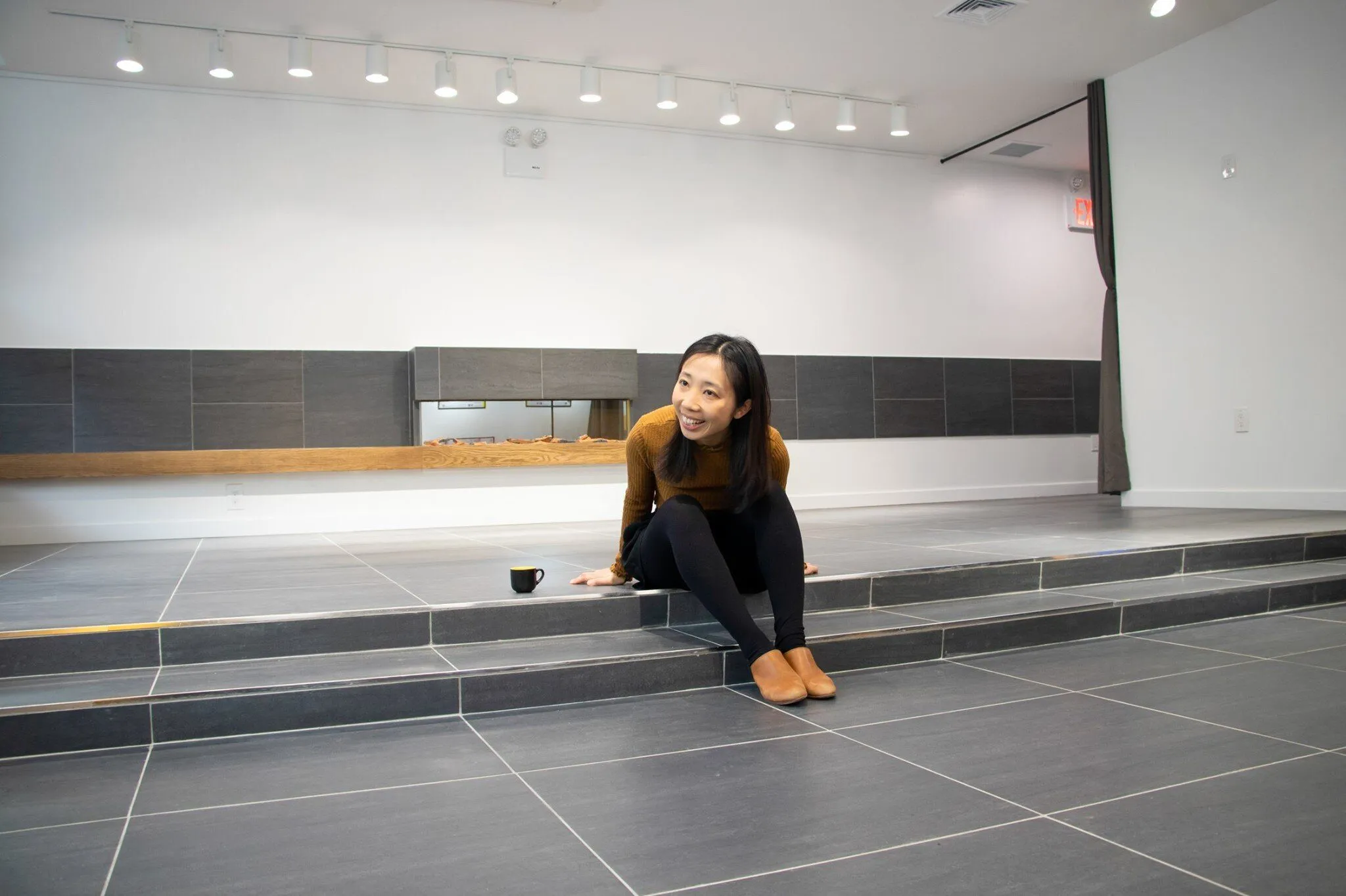 A woman sits on a raised, two-step platform with a long built-in modern fireplace behind it, beneath a row of track lights.
