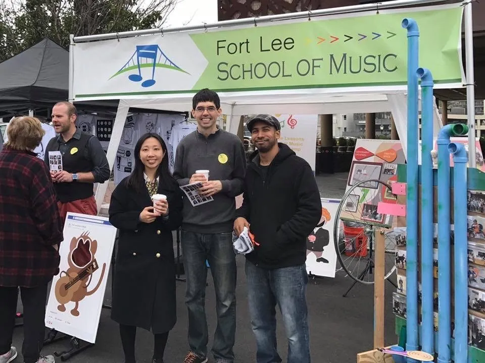 Staff stand in front of a Fort Lee School of Music booth with a large green-and-white logo banner at an outdoor street festival.
