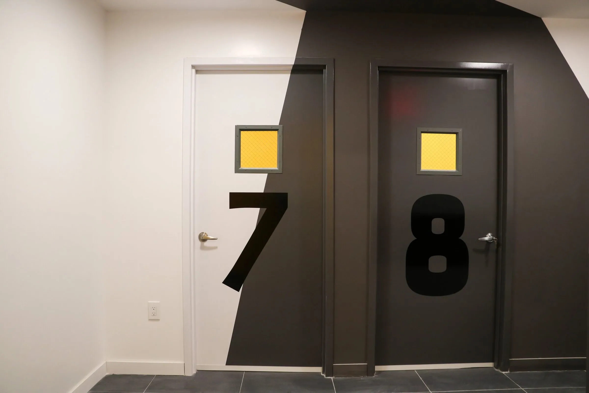 Two studio doors marked with oversized black numerals 7 and 8, small yellow square windows, and a charcoal diagonal painted across the wall.